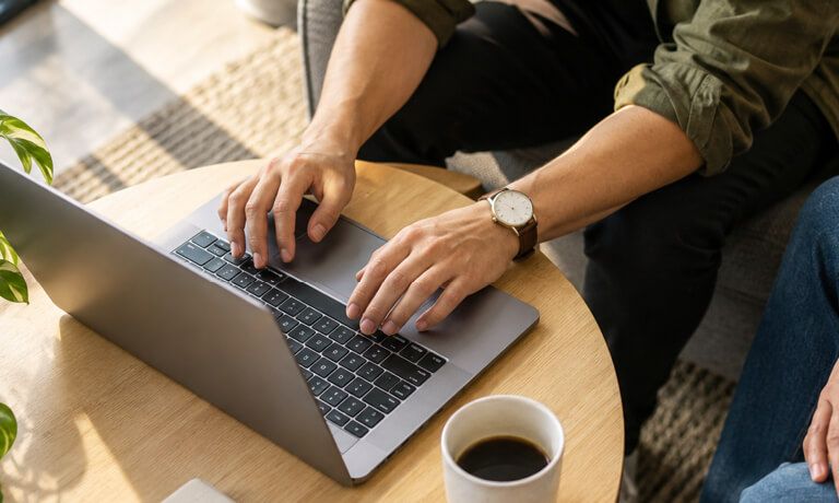 Hands typing on a laptop at a wooden table next to a cup of coffee.