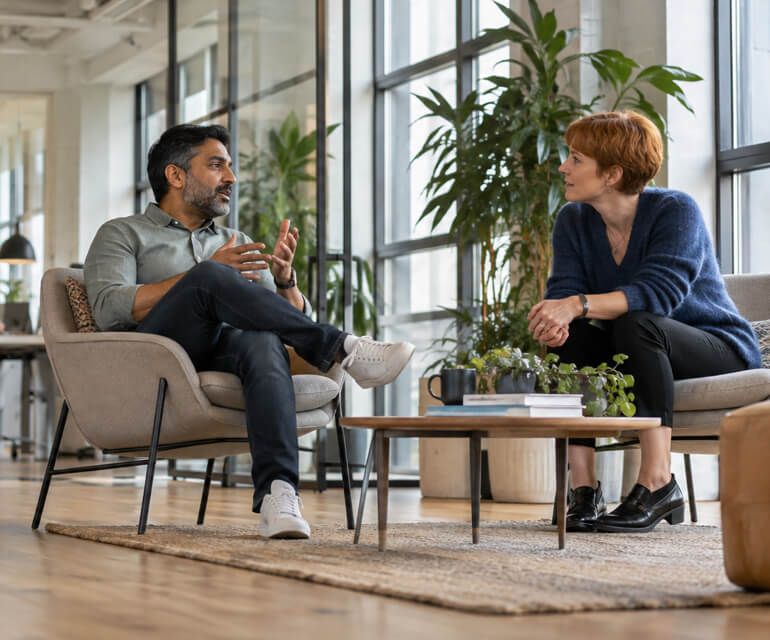 Two colleagues in armchairs talking in a bright office lounge.