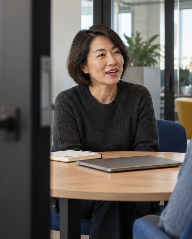 A woman at a meeting table with a laptop, smiling mid-conversation.