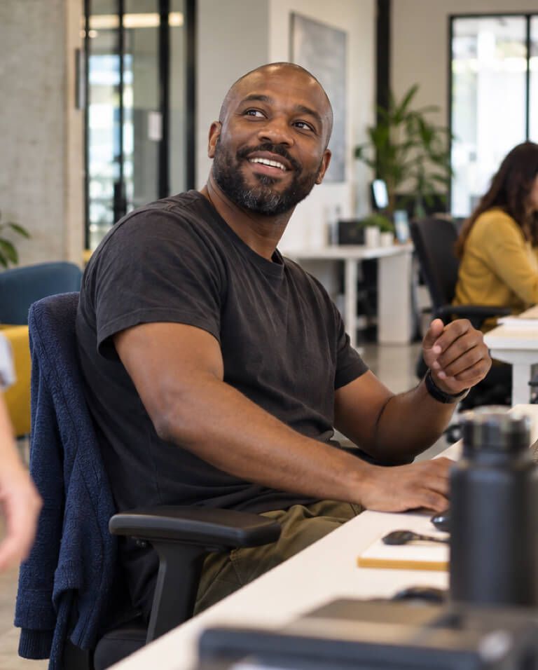 A smiling man at his desk, colleagues working in the background.